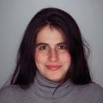 smiling woman with long, dark hair and a gray turtleneck standing against a solid gray background