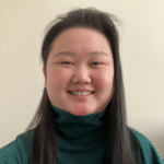 smiling woman with long dark-colored hair wearing a greenish turtleneck seated against a beige background