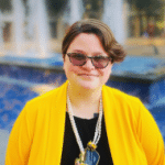 profile photo of woman with short brown hair, dark glasses, and wearing a yellow sweater over a black shirt standing outside