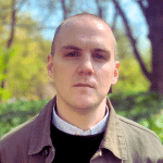 man with short hair wearing a green collared shirt, dark colored sweater, and light colored collared shirt standing outdoors