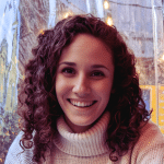 smiling woman with curly brown hair wearing a white turtleneck sweater sitting outdoors