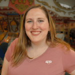 smiling woman with reddish hair and wearing a pink shirt standing in front of a carousel