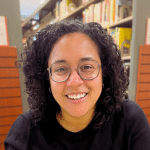 smiling woman with shoulder-length, dark curly hair and round-rimmed glasses, wearing a black shirt and sitting in a library