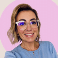 smiling woman with short blonde hair and light-rimmed glasses wearing a blue sweater and standing in front of a pink and beige background