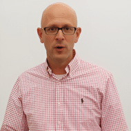 man with glasses and pink collared shirt standing against a grayish background