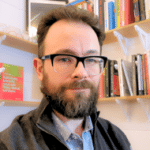 profile photo of man with short dark hair, dark-rimmed glasses, and a beard wearing a gray sweater and collared shirt seated in front of a bookshelf