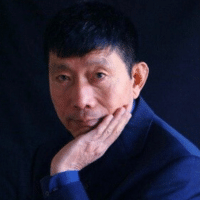 headshot of man with dark hair in blue sweater resting chin in hand sitting against a dark background