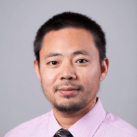 man with short dark hair and goatee wearing a pink shirt and dark tie standing against a grayish background